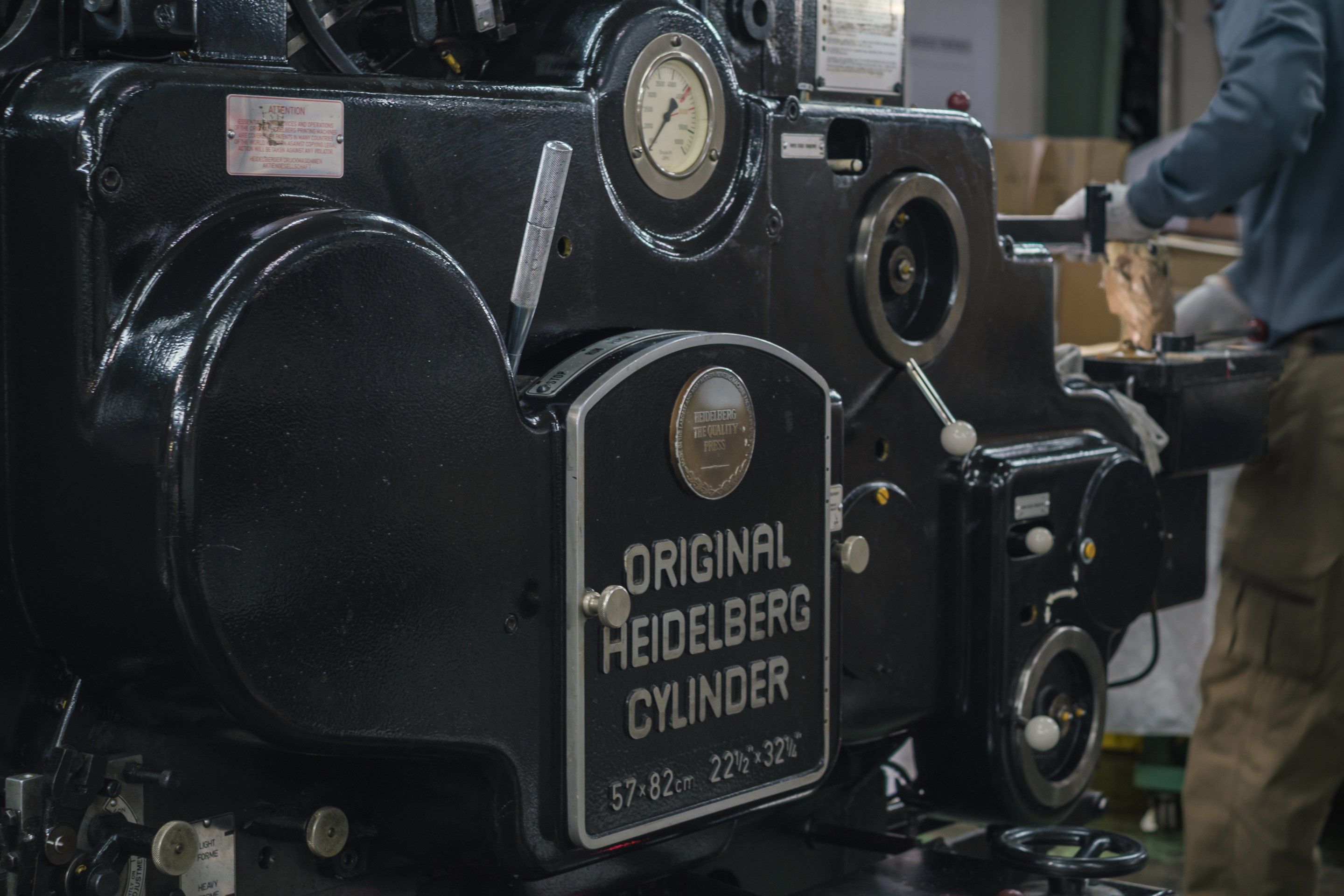 黒い印刷機の側面の銘板。「ORIGINAL HEIDELBERG CYLINDER」と浮き彫りになっている。奥で作業着の人が機械に手を伸ばしている