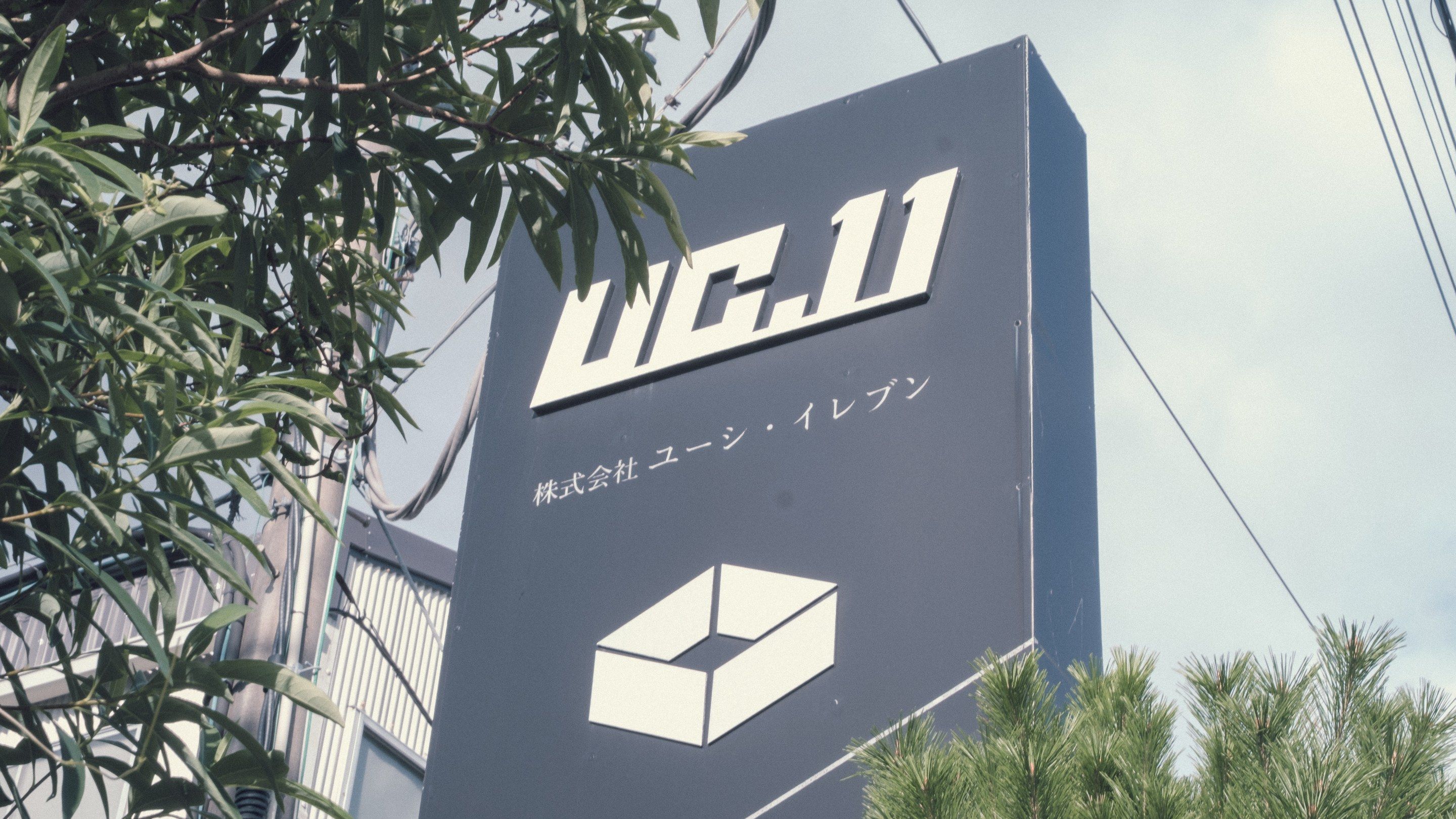 濃い灰色の看板に白い字で「UC.11」「株式会社ユーシ・イレブン」とあり、その下に箱を上から見た形の印がある。左に木の葉、右下に松の枝